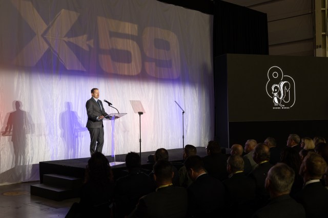 A man speaks at a podium. In the immediate background, the X-59 sits unseen behind a large curtain, moments away from its public unveiling.