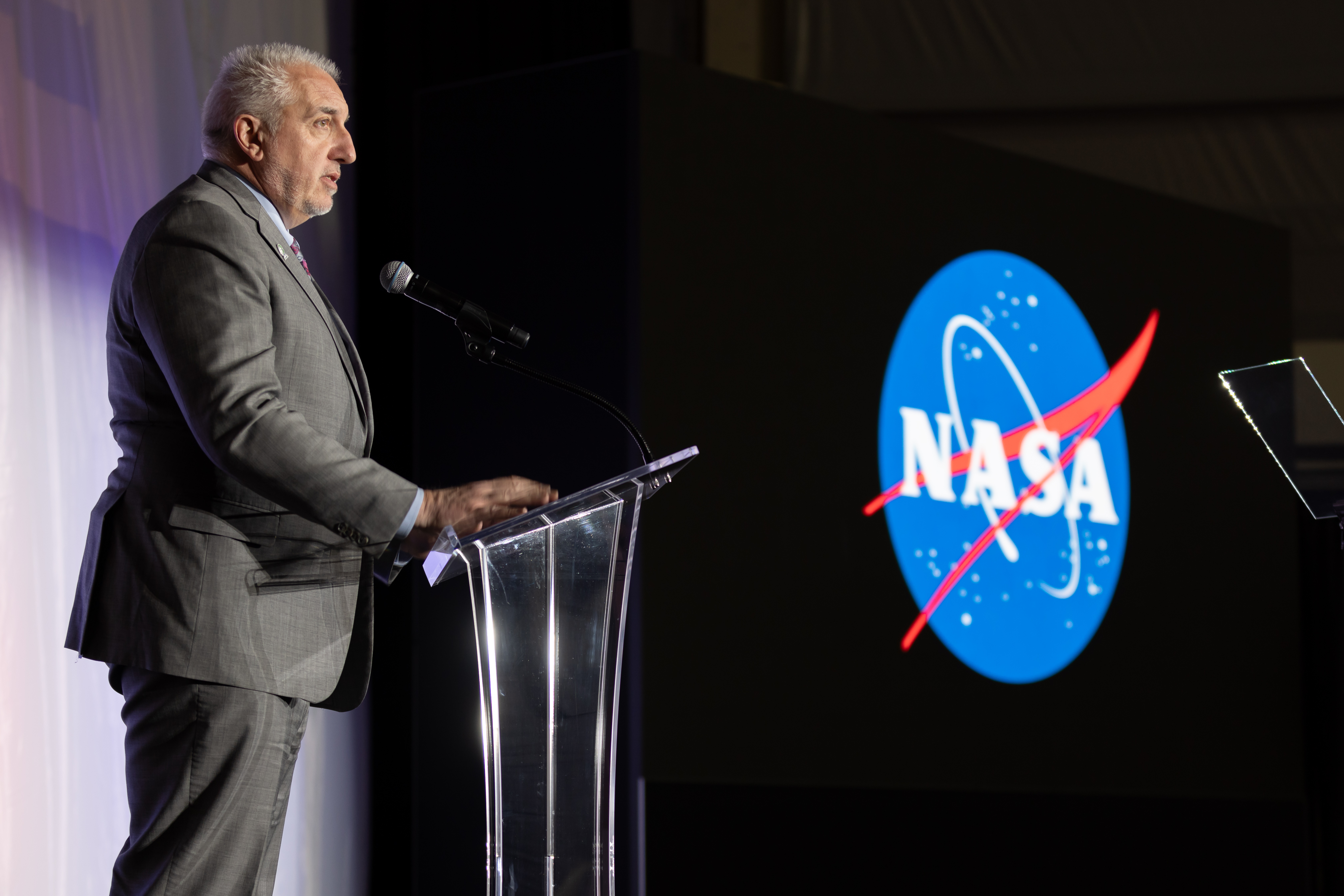 A man speaks at a podium. In the immediate background, the X-59 sits unseen behind a large curtain, moments away from its public unveiling.