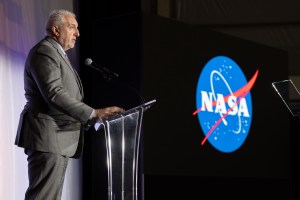 A man speaks at a podium. In the immediate background, the X-59 sits unseen behind a large curtain, moments away from its public unveiling.