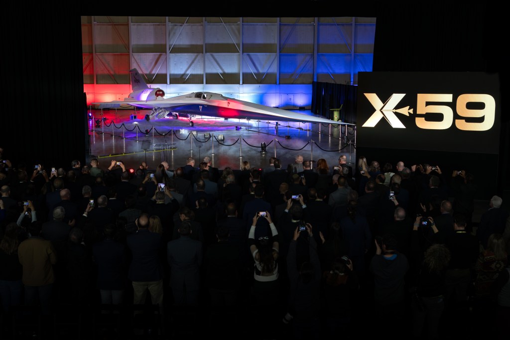A large crowd stands before NASA's X-59 aircraft, many holding mobile devices over their heads.