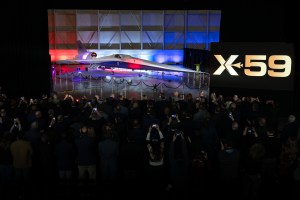 A large crowd stands before NASA's X-59 aircraft, many holding mobile devices over their heads.