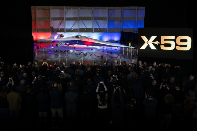 A large crowd stands before NASA's X-59 aircraft, many holding mobile devices over their heads.
