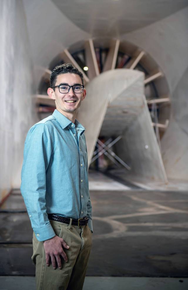 Young engineer wearing glasses and a blue shirt smiles at camera in front of a large fan inside the Icing Research Tunnel at NASA Glenn. 