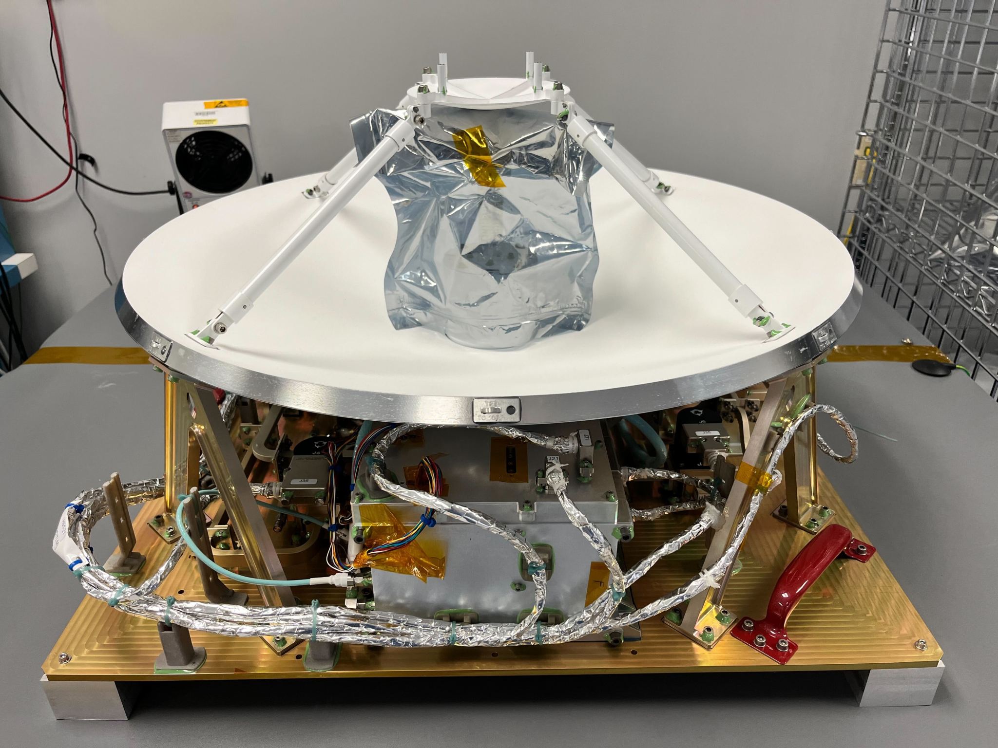 The Polylingual Experimental Terminal is the focus of this photograph. We see a white antenna dish, approximately 0.6-meters in size, facing the ceiling, sitting on a golden platform. Silver wires resembling tinfoil are shown protruding beneath the antenna dish. The terminal sits on top of a grey table inside a white laboratory.