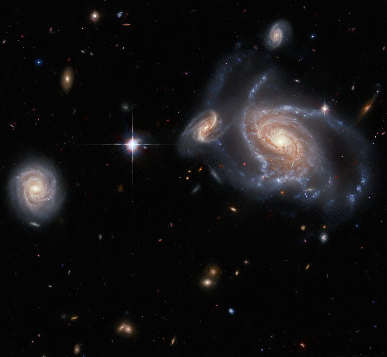 Against the blackness of space, one very large spiral galaxy and three smaller spiral galaxies are prominently visible amidst the other colorful specks of light.