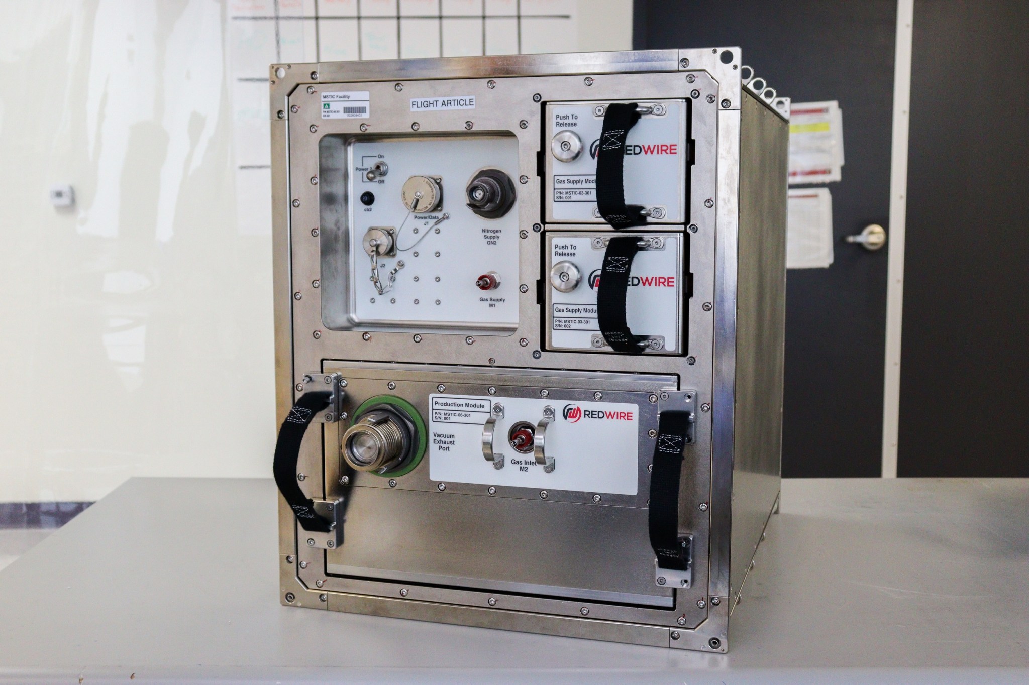 A sturdy silver box larger than a microwave oven has a large panel across the bottom labeled “Vacuum Exhaust Port,” two panels in the upper right labeled ‘Gas Supply Module,” and an inset on the upper left with several connectors. The panels have black handles.