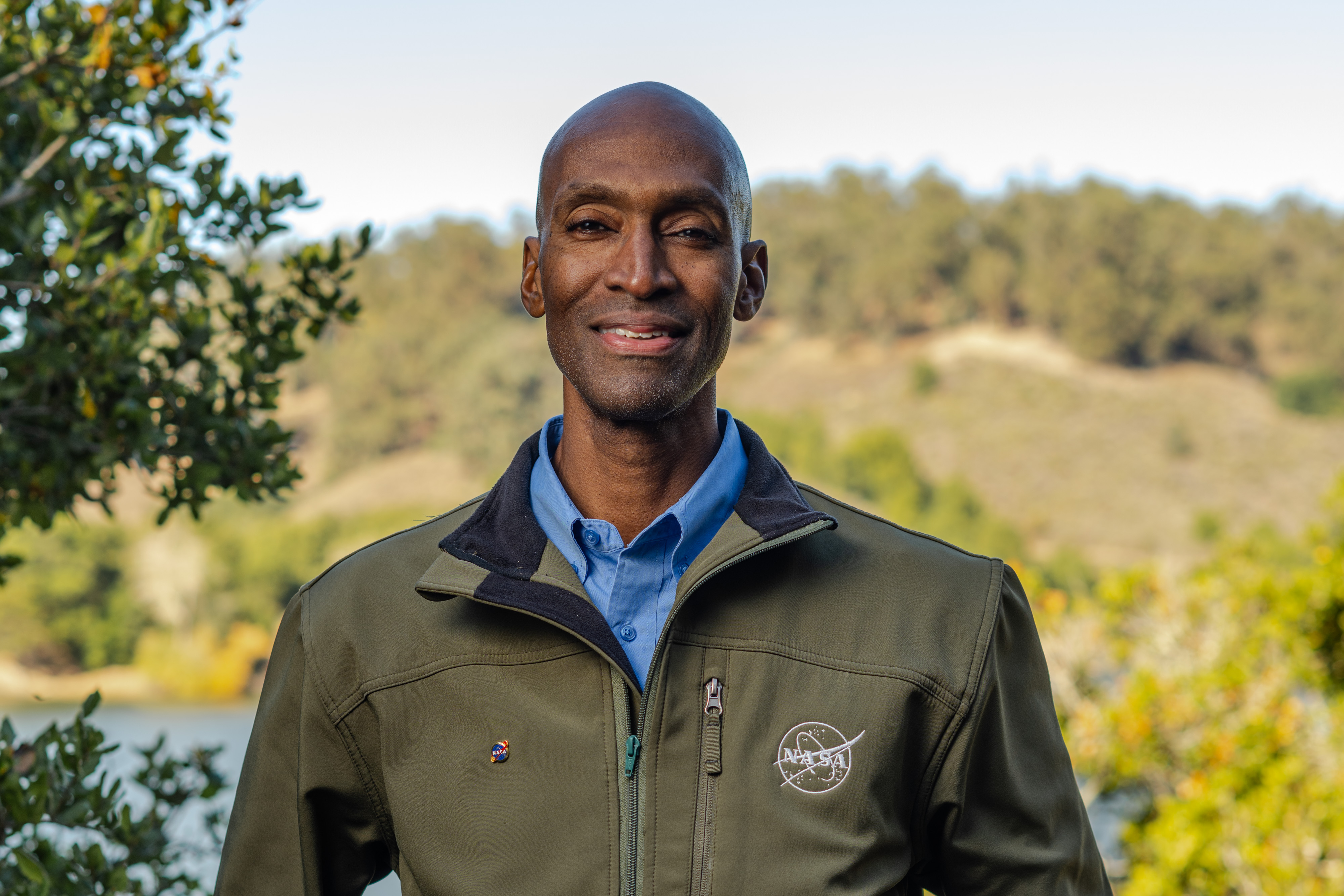 Rodney Martin, wearing an olive-colored NASA jacket and a blue dress shirt underneath, smiles at the camera. He is standing in the foreground with Lake Chabot Park in the background.