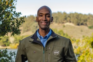 Rodney Martin, wearing an olive-colored NASA jacket and a blue dress shirt underneath, smiles at the camera. He is standing in the foreground with Lake Chabot Park in the background.