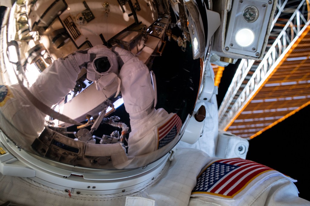 A spacewalker's spacesuit gloves and camera are reflected in the helmet visor in this "space-selfie" taken during a six-hour and seven-minute spacewalk.