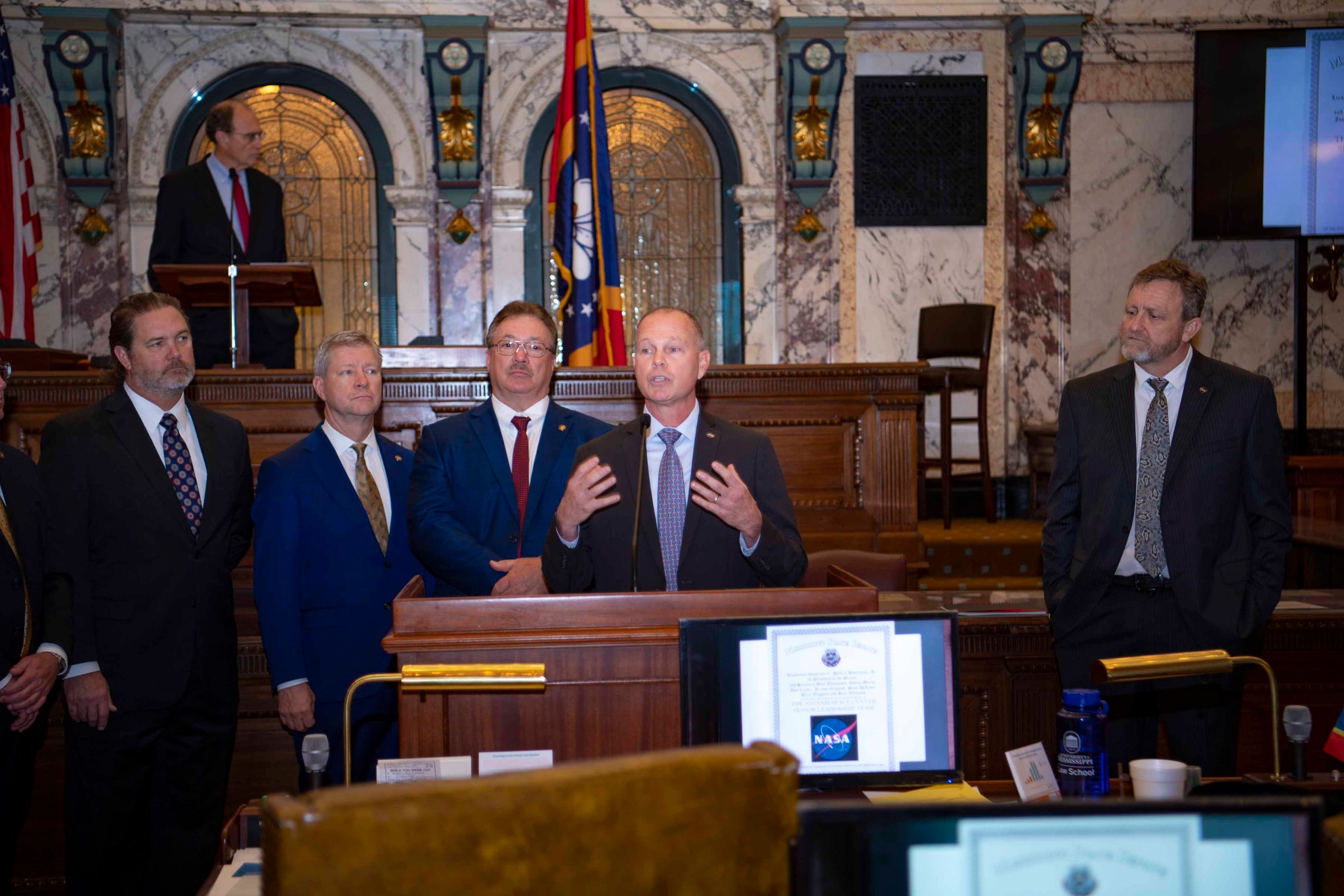 man speaks from behind lectern with other leadership members nearby