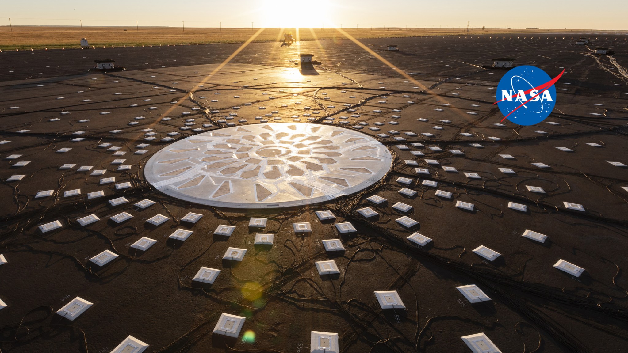 Large white circular solar panel on a black ground surface surrounded by many smaller white square solar panels with sunshine beaming in from the top.