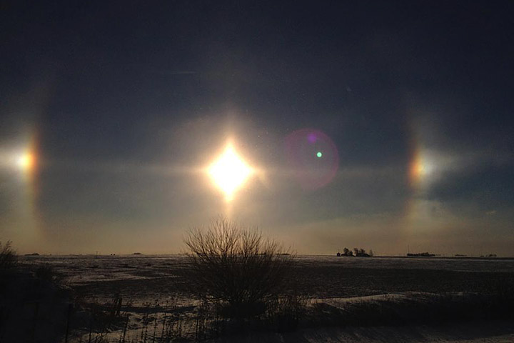 The focal point of the image is the bright sun, centered. Two reflections of the sun are seen to the left and right of the main bright spot. Below the sun is a grassy landscape with a bush directly below the sun. The sky is a dark blue color.