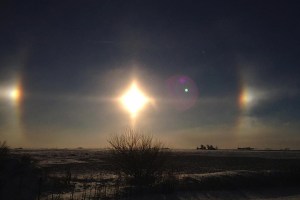 The focal point of the image is the bright sun, centered. Two reflections of the sun are seen to the left and right of the main bright spot. Below the sun is a grassy landscape with a bush directly below the sun. The sky is a dark blue color.