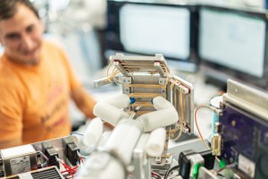 A white robot about the size of a hand with two large fingers that end in metal probes faces a metal three-part panel with rubber bands stretched across it, which the robot is cutting. A lab worker and computer screens are blurred in the background.