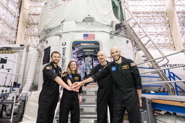 Four astronauts in NASA jumpsuits standing in front of a space capsule