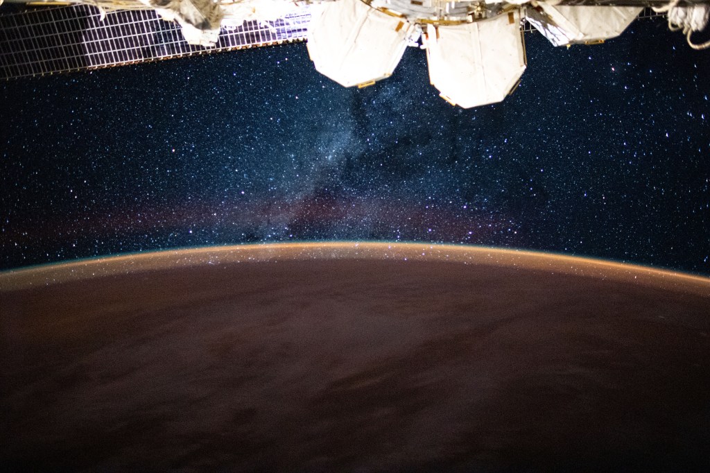 This long-exposure photograph from the International Space Station was taken during an orbital night period and reveals the Milky Way glittering above a bright but exaggerated atmospheric glow blanketing the Earth's horizon.