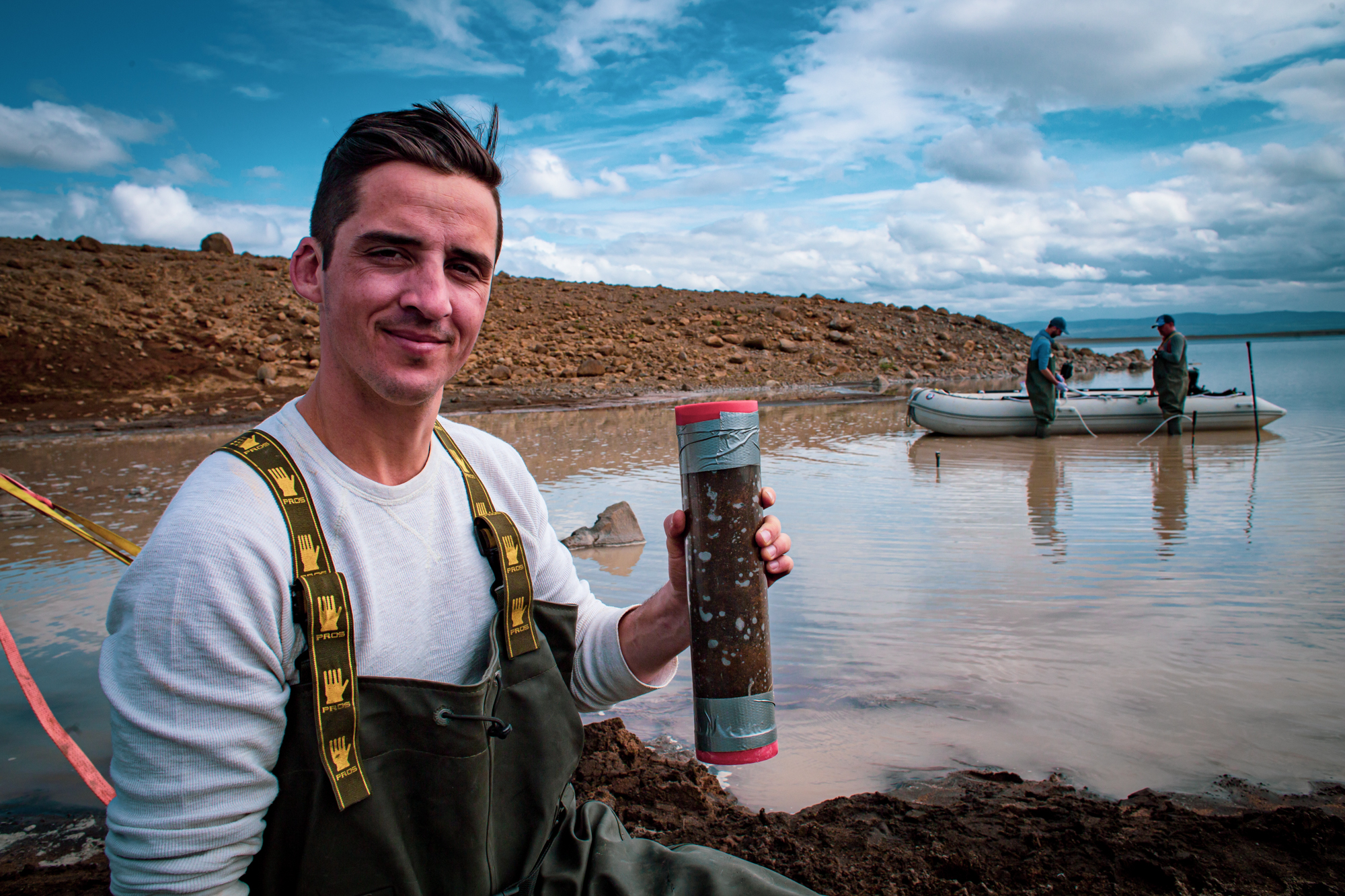 Michael Thorpe, a man with dark hair, poses with a tube of sediment collected from a nearby body of water. He wears a gray sweatshirt underneath dark green waders, or waterproof overalls, and kneels next to a brown lake with clumpy dirt banks in the foreground and background. Two more scientists in waders stand next to a small, flat white boat. The sky is bright blue and mostly covered with puffy clouds.