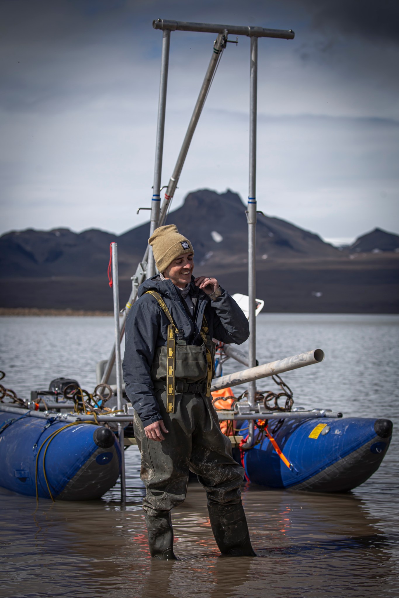 Dr. Michael Thorpe laughs and adjusts his coat collar as he stands in ankle-deep water. He wears a gold stocking cap and navy blue winter coat underneath dark green waders, or waterproof overalls. He stands in front of a blue pontoon boat covered with straps, loops, and equipment. Dark brown, rocky mountains are visible in the background and the sky is a heavy, cloudy gray.
