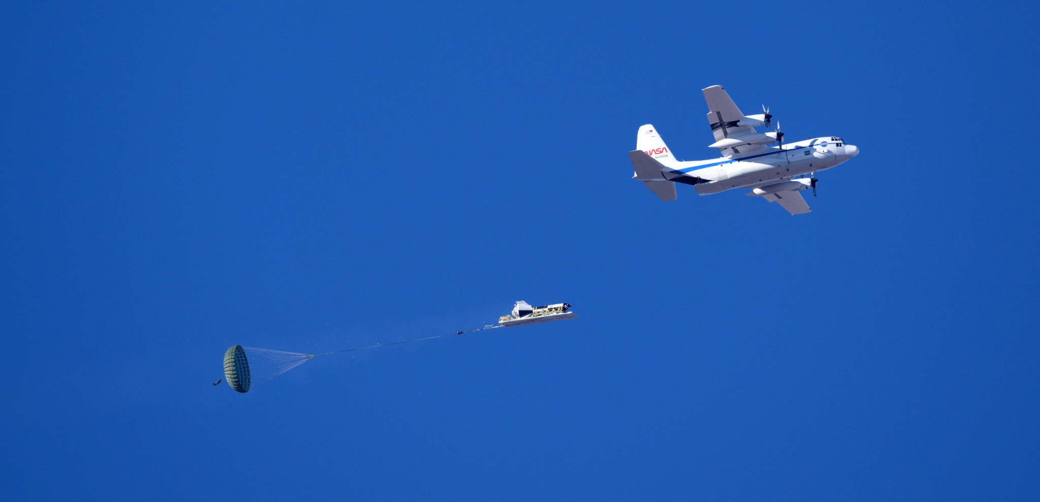 A NASA C-130 cargo aircraft, flying in clear skies, has dropped a test vehicle from its open cargo door. The test vehicle is shaped like a dart and has a large ring-like parachute attached to it.