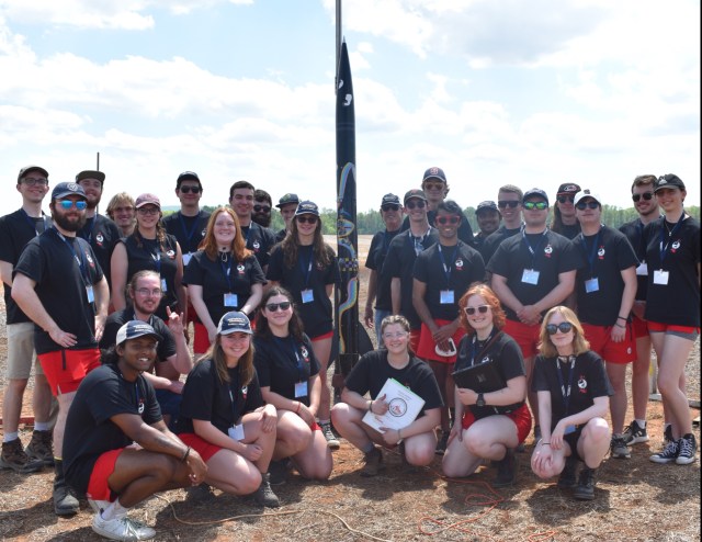 A group of students in black t-shirts and red shorts pose around a student created rocket.