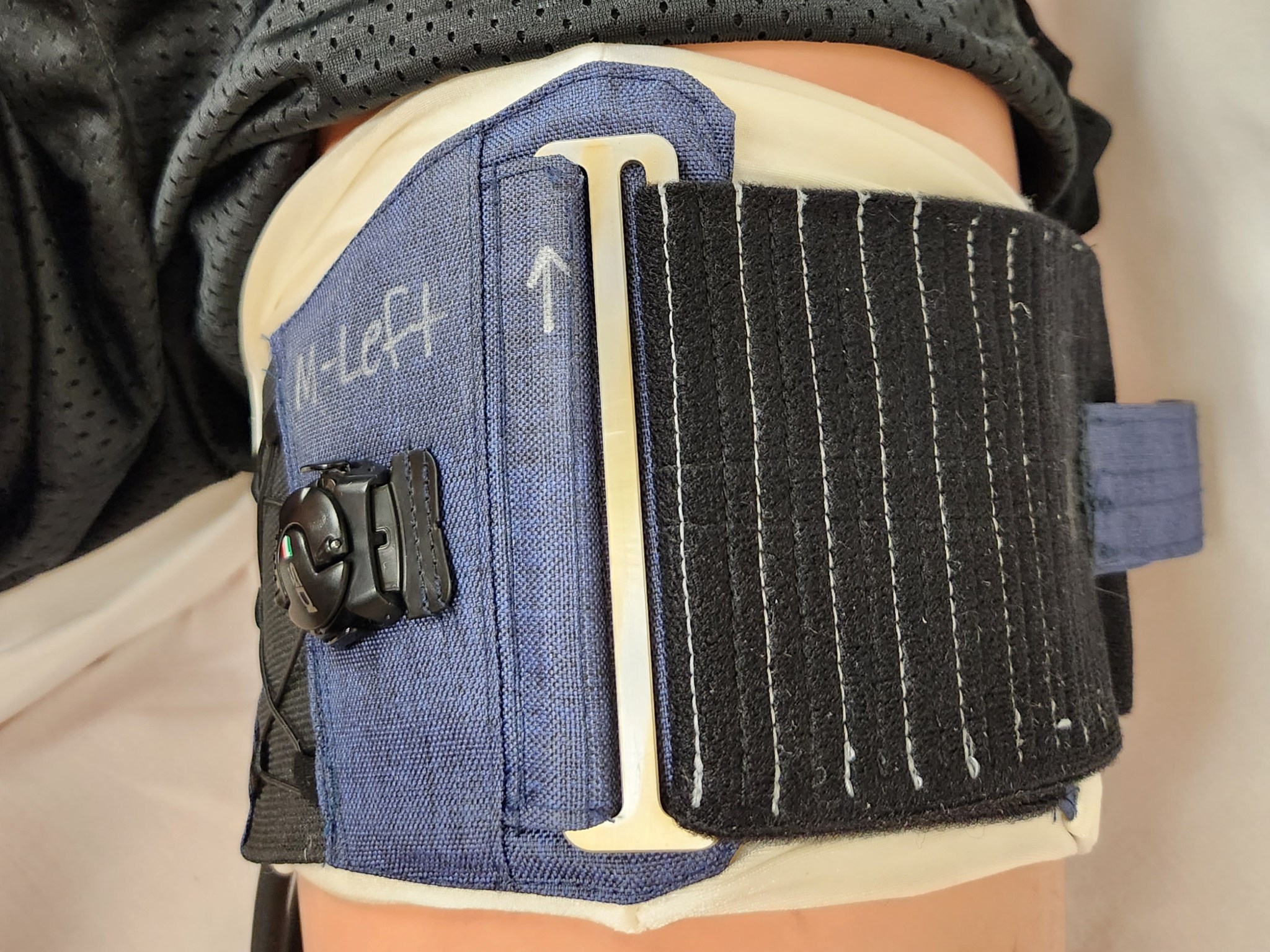 A person wearing black shorts lies on a white sheet with a thigh cuff around their upper leg. The cuff is blue with a black Velcro attachment and is labeled “left.”
