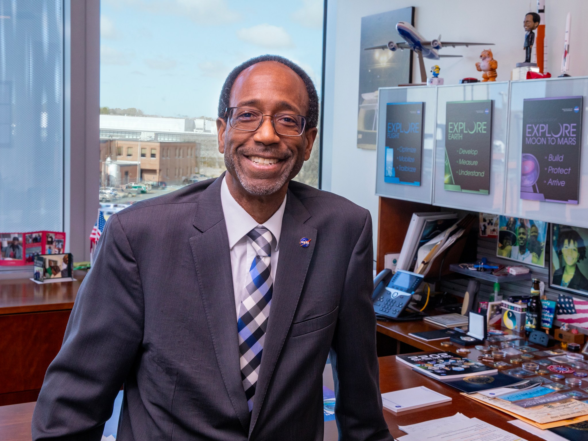 This is a photo of Clayton P. Turner, Director of NASA’s Langley Research Center in Hampton, Virginia. Clayton is wearing a suit and tie and sits on a desk. On his desk are papers, pens and pencils, medals, photos, an American flag, a model plane, and other assorted memorabilia. Buildings can be seen through a window in the background of the photo.