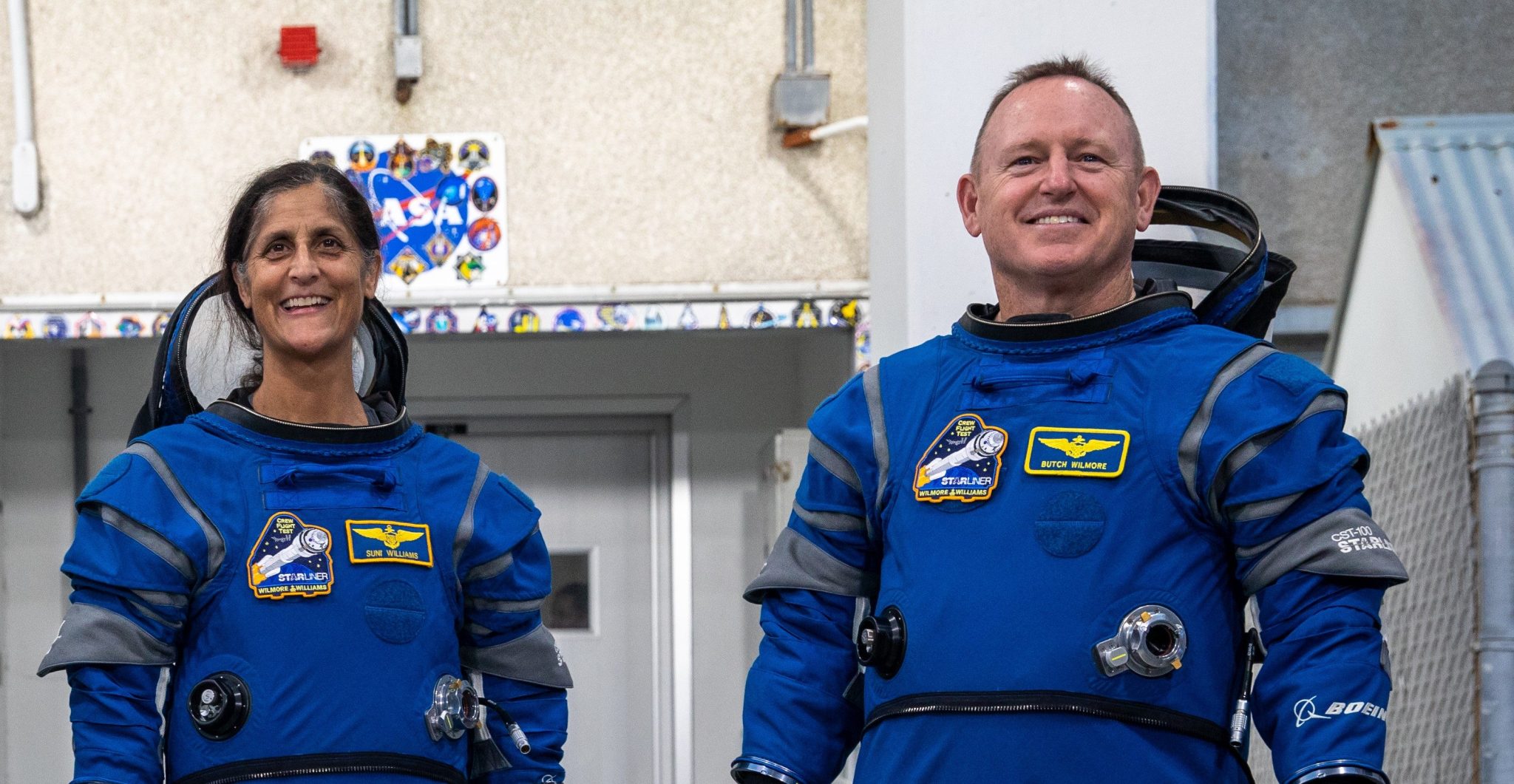 NASA astronauts Suni Williams and Barry “Butch” Wilmore, exit the Astronaut Crew Quarters at NASA’s Kennedy Space Center in Florida. They are both wearing blue flight suits with the mission patch and name patches visible on the front, as well as white shoe covers on their feet.