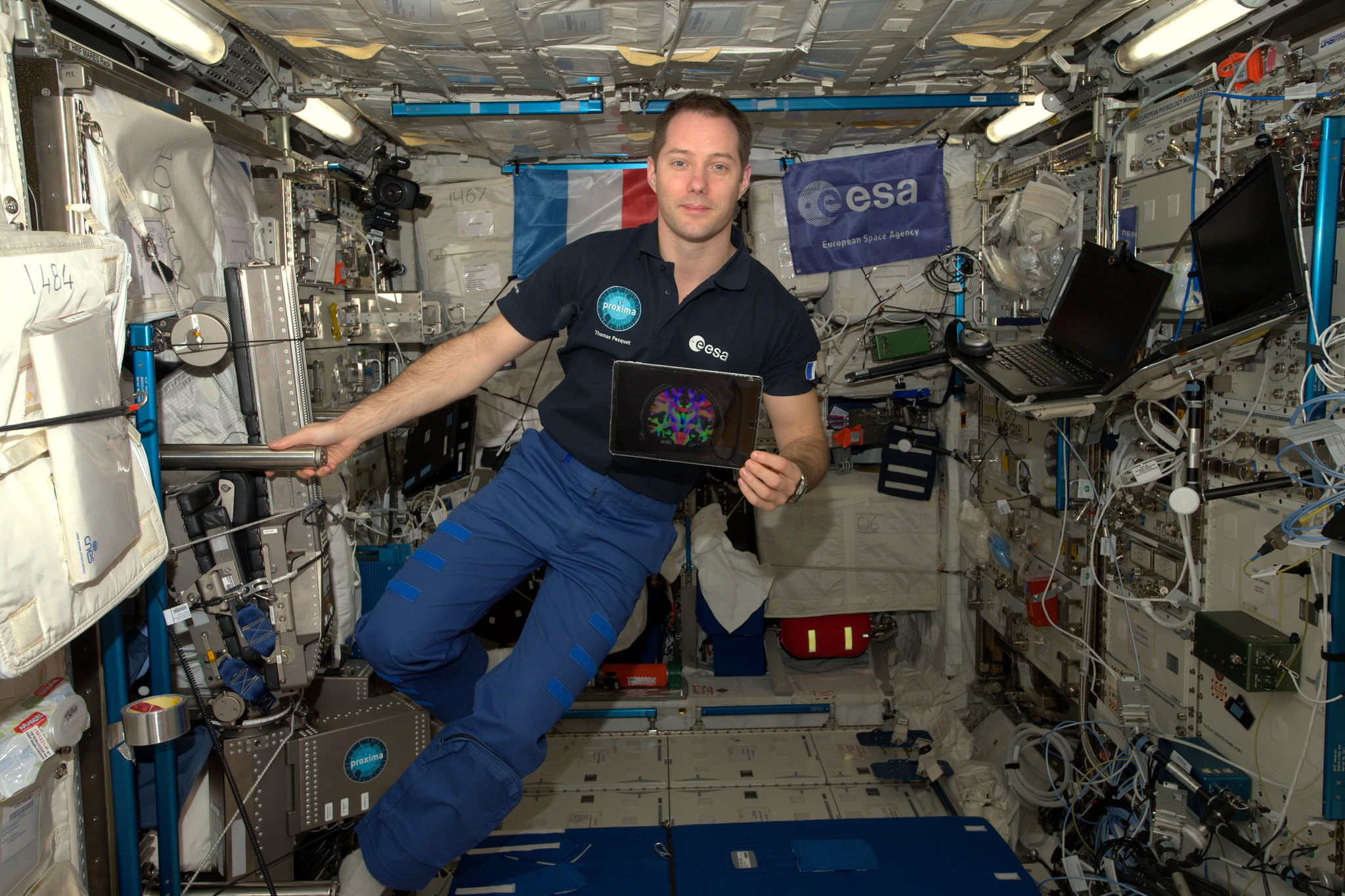 Pesquet is wearing a dark polo shirt with the ESA logo and a pair of light blue pants. He is facing the camera and holding on to a bar extending from a wall of the space station with his right hand. In his left hand is a tablet with a multi-colored image on it. There are ESA and French flags on the station wall behind him and laptops, equipment, and cords covering the wall to his left.