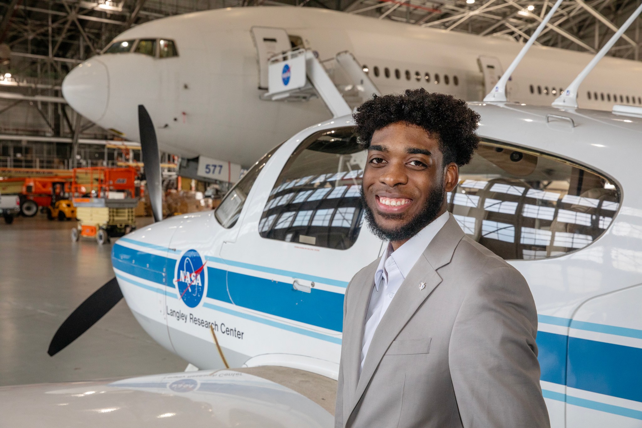 This is a photo of Brandon Sells, an aerospace engineer with the Aeronautics Systems Analysis Branch (ASAB) of the Systems Analysis and Concepts Directorate (SACD) at NASA Langley Research Center. Brandon is standing in front of a small white and blue plane with a propellor. A larger white plane can be seen in the background of the photo.