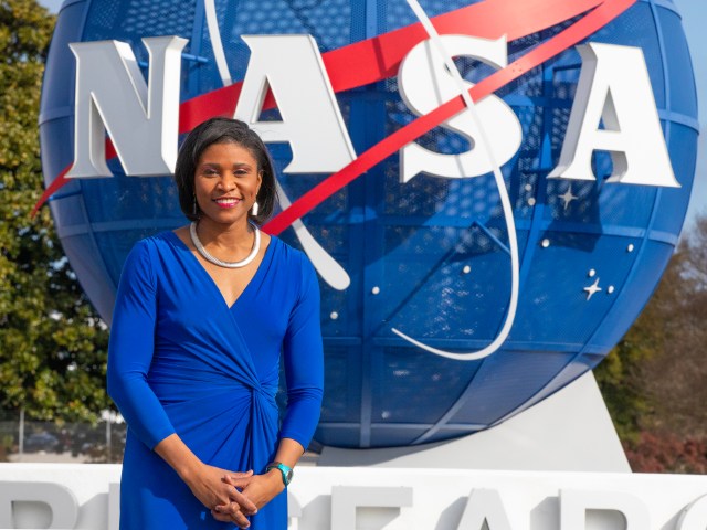 This is a photo of Brittny McGraw, News Chief in the Office of Communications at NASA’s Langley Research Center. She joined NASA Langley in September 2023, after a 20-year career as an award-winning broadcast journalist. Brittny is wearing a blue dress and is standing in front of the large welcome sign at NASA Langley Research Center, which features a 3-D sculpture of the NASA logo. Trees can be seen in the background of the image.