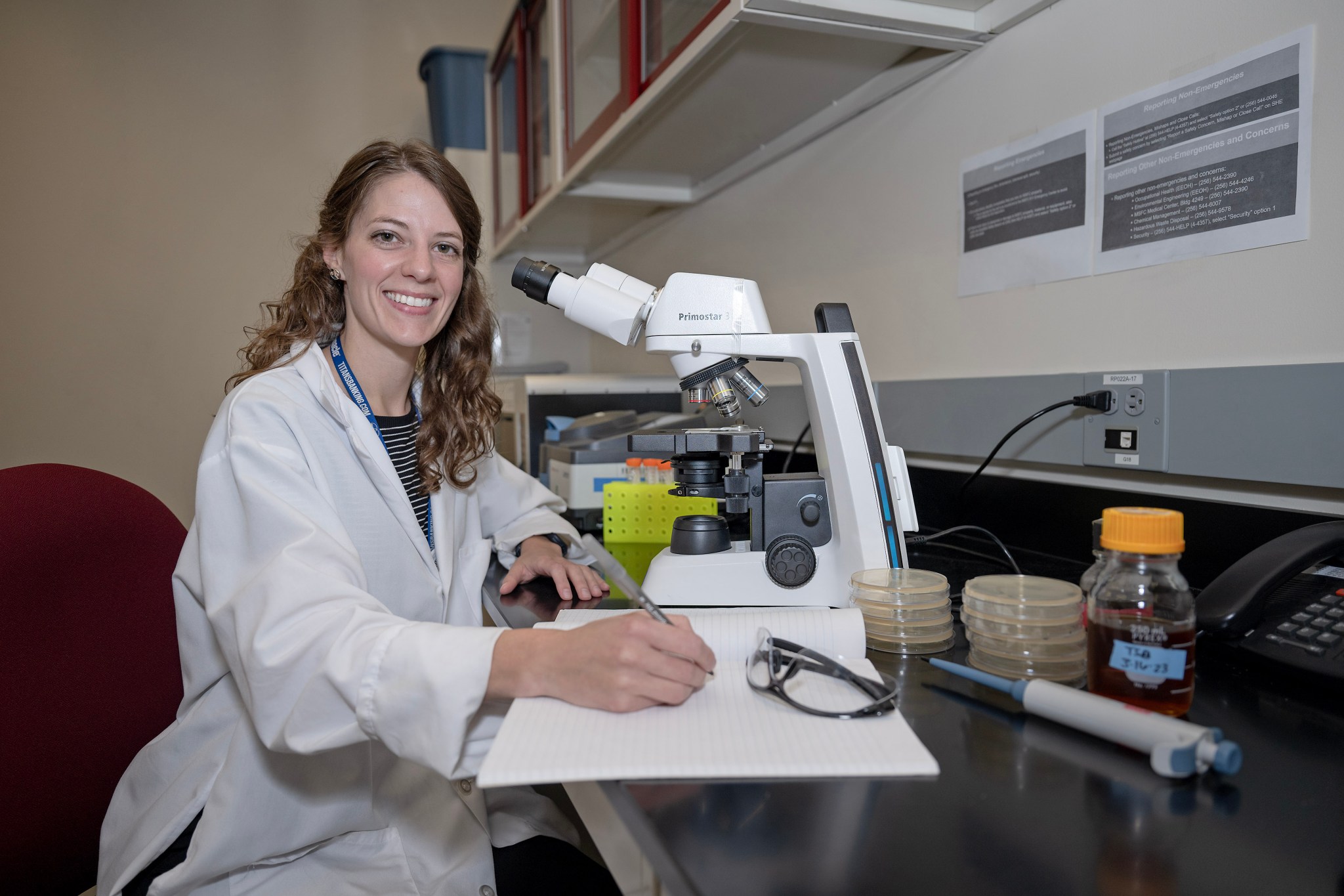 Planetary Protection microbiologist Chelsi Cassilly sits at a microscope in a white lab coat smiling at the camera.