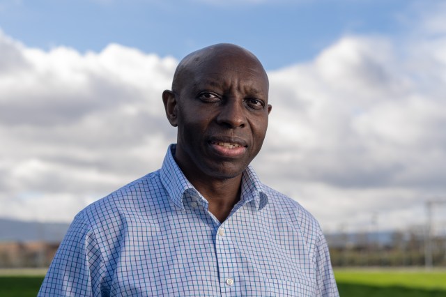 Charles Gatebe, wearing a blue checkered dress shirt, looks at the camera against a background of blue skies, white fluffy clouds, and green grass.