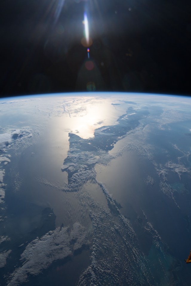 This view of Cuba looks from east to west and reaches the cities of Santiago de Cuba on its west coast and Havana on its east coast. The sun's glint is reflected off the Caribbean Sea on Cuba's southern coast.