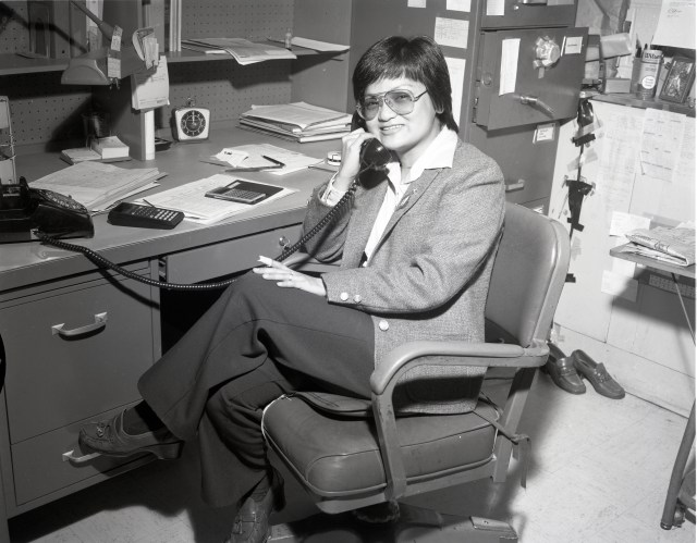 In this black-and-white photo, Doris Britton holds a corded telephone receiver up to her ear. She is wearing professional attire and sits at a desk strewn with papers and calculators. She smiles at the camera. 