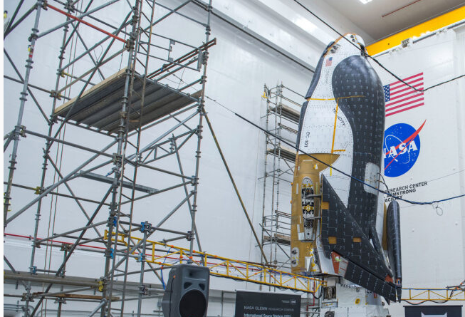 Sierra Space’s Dream Chaser spaceplane and its Shooting Star cargo module stacked inside NASA’s Neil Armstrong Test Facility in Sandusky, Ohio. The spaceplane is black, white, and dark orange, and its wings are folded up. A NASA meatball and an American flag can be seen on the wall behind the spaceplane. Speakers, banners, and a podium are set up in the bright room for a media event.