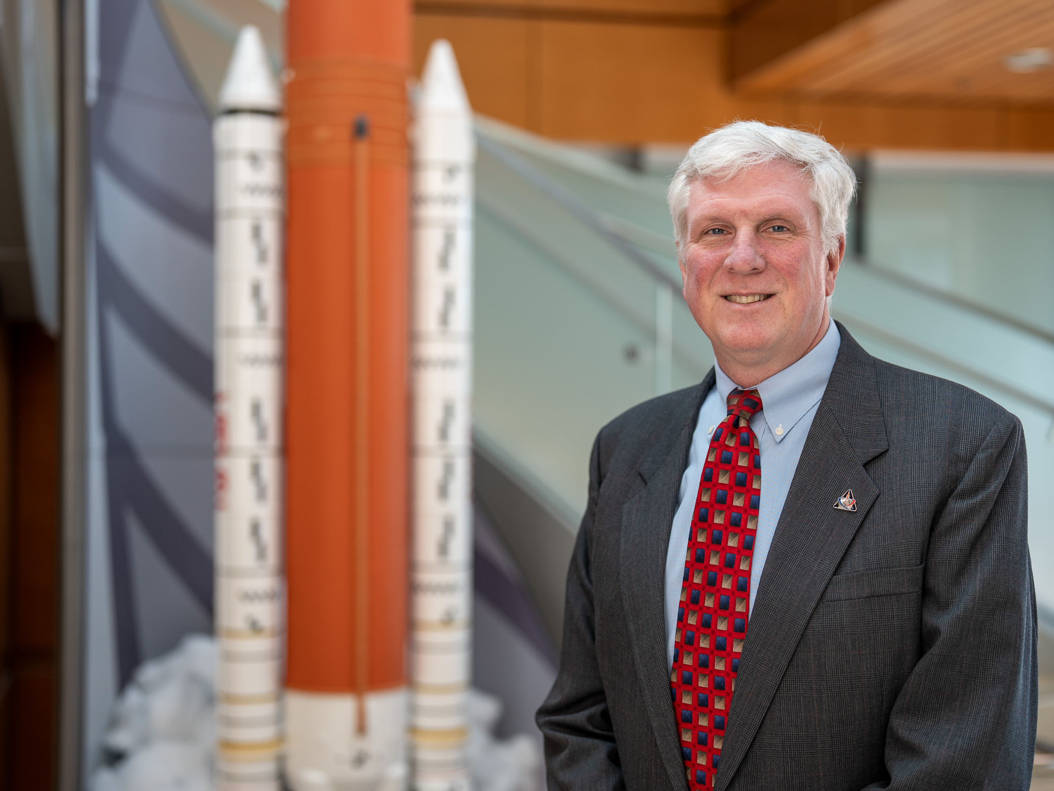 Matthew Ramsey poses for a photo in front of a model of the SLS rocket. He is wearing a grey suit, light blue shift and a red and black patterned tie.