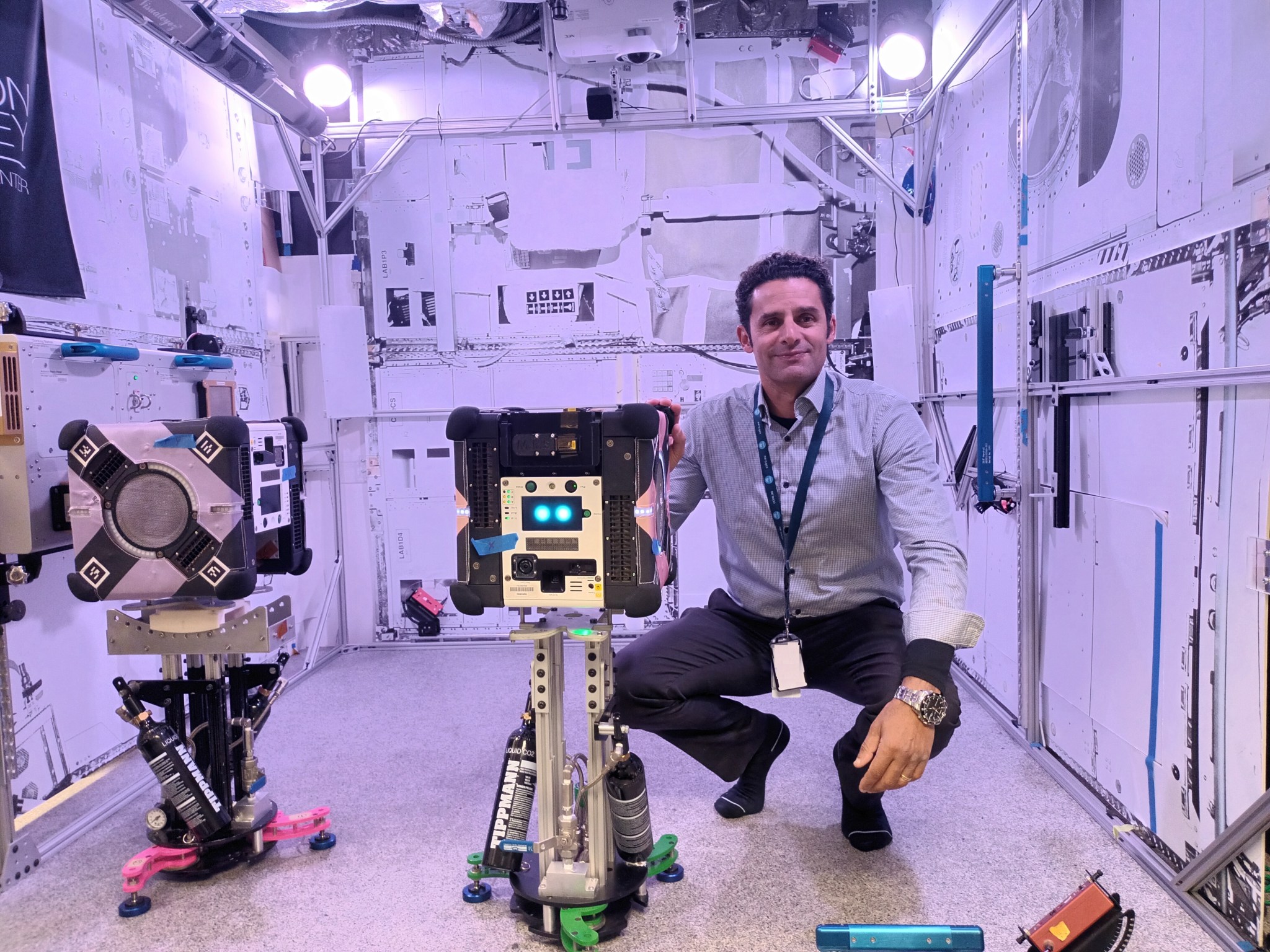 Inside a large white test container, Elmouttie crouches next to a carry-on suitcase-sized box with two camera lenses visible on the top front of the box and the Astrobee robot’s white control panel below it with two round glowing lights.