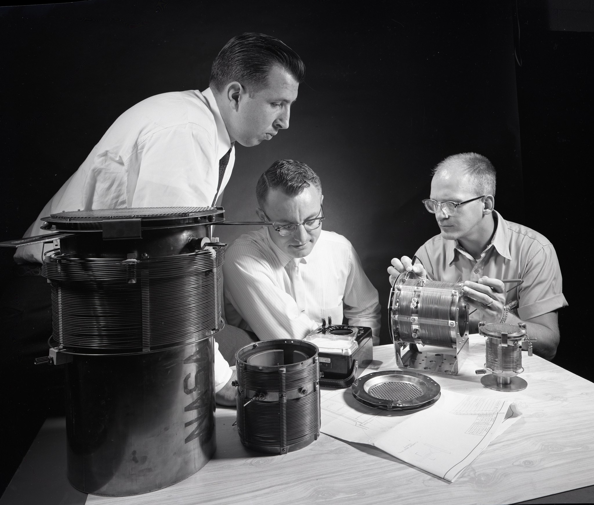 Three men working with components at table.