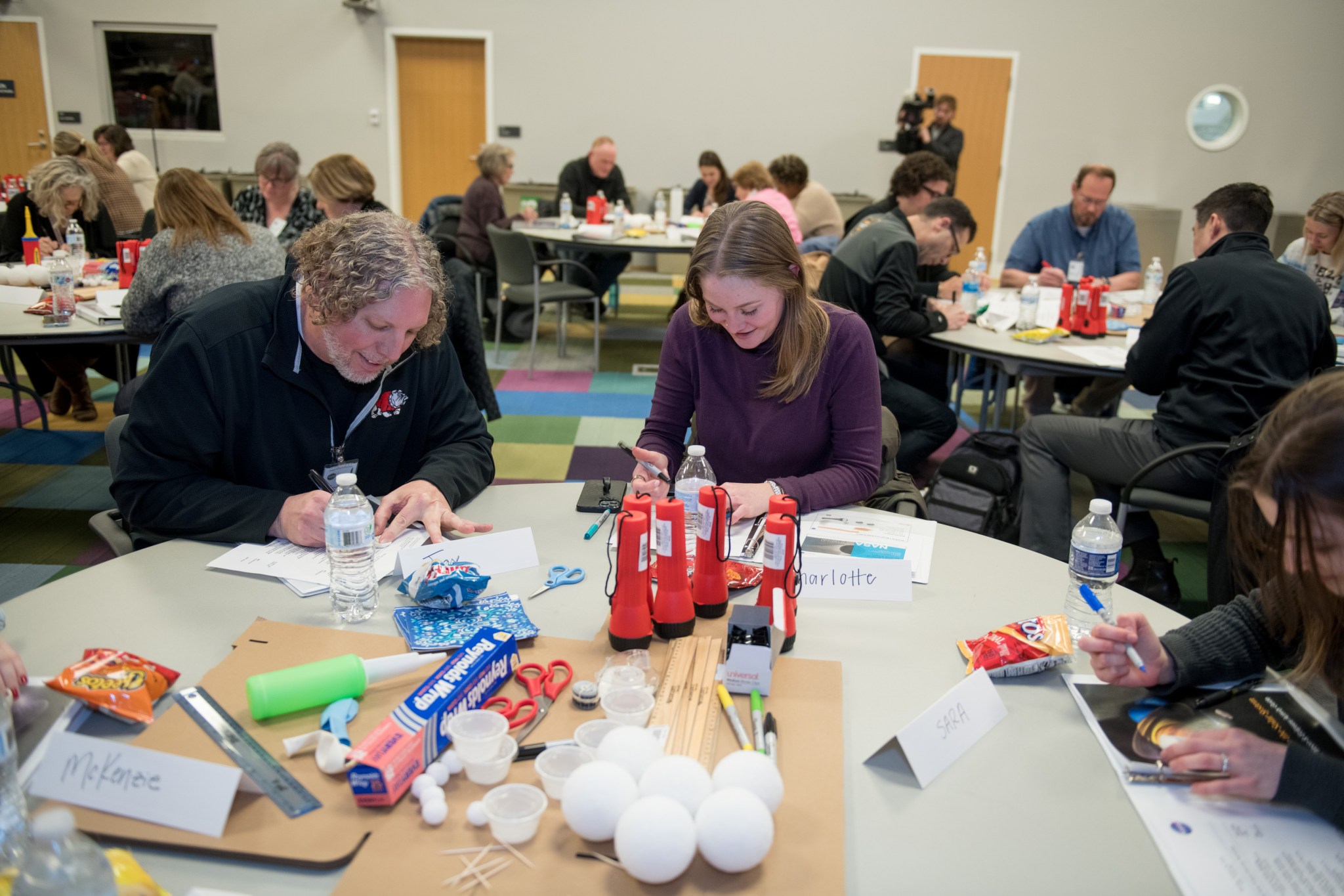 Several tables contain flashlights, rulers, scissors, tinfoil, and foam balls. Educators read instructions before constructing box pinhole projectors.