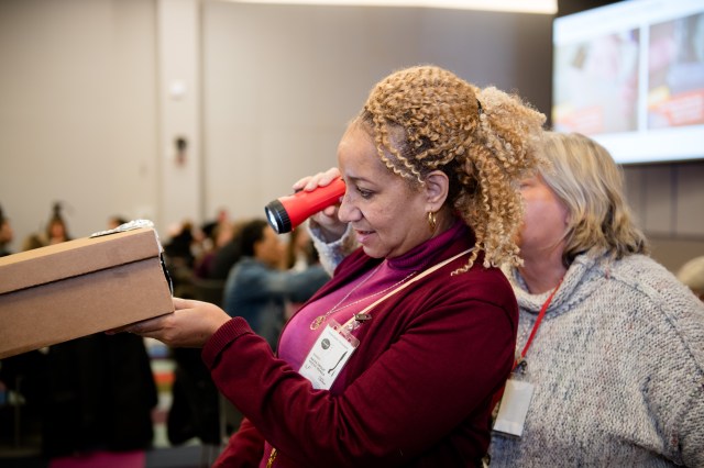 Educators test construction box pinhole projectors for solar eclipse viewing. An educator looks into a small hole in a shoebox as another educator stands behind her and holds a flashlight facing the box.
