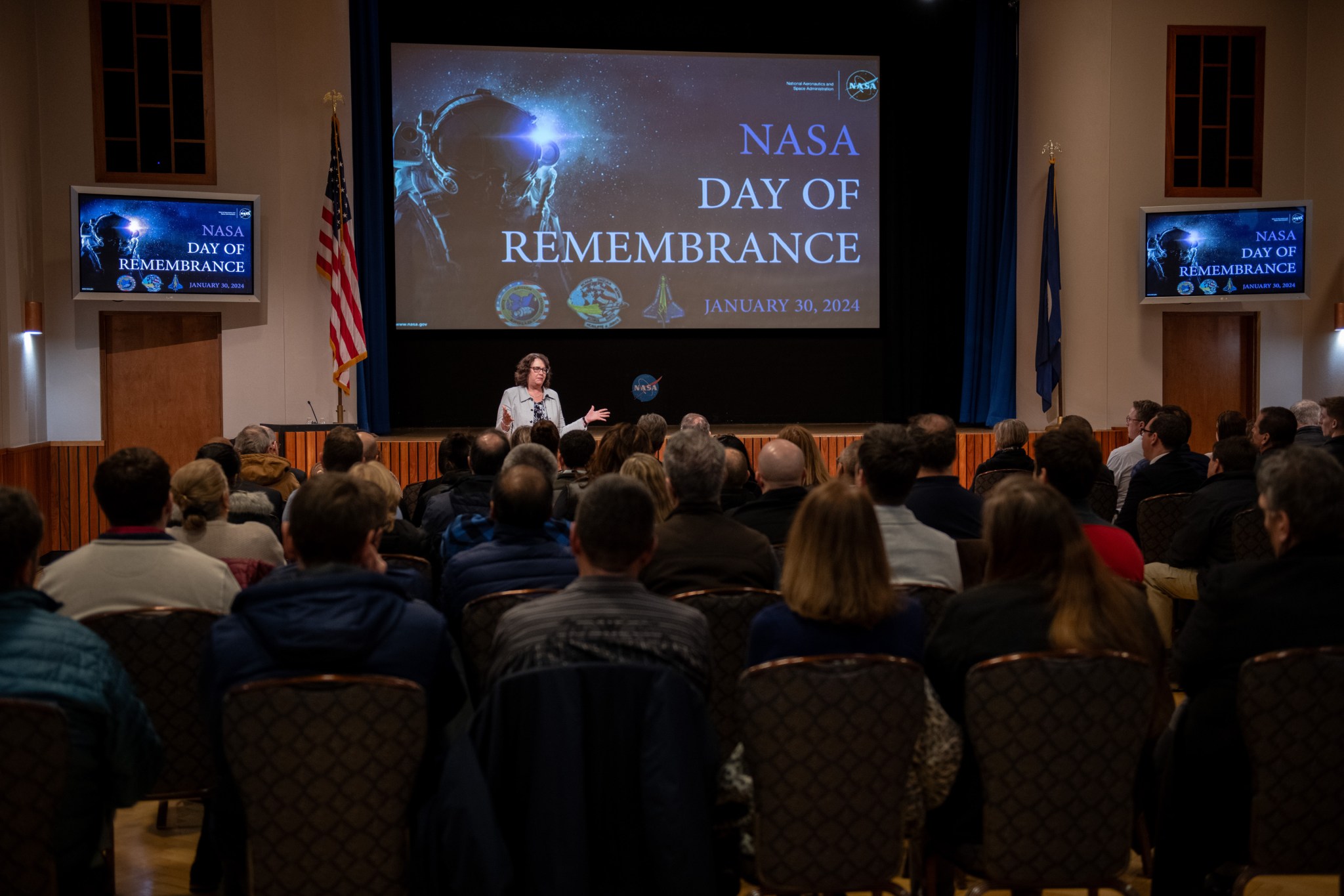 NASA Glenn Deputy Director Dawn Schaible stands at the front of an auditorium and addresses employees who are seated in the audience. A screen with the words, “NASA Day of Remembrance,” with three mission patches are behind her. Two smaller screens with the same information are at each side of the front of the room.