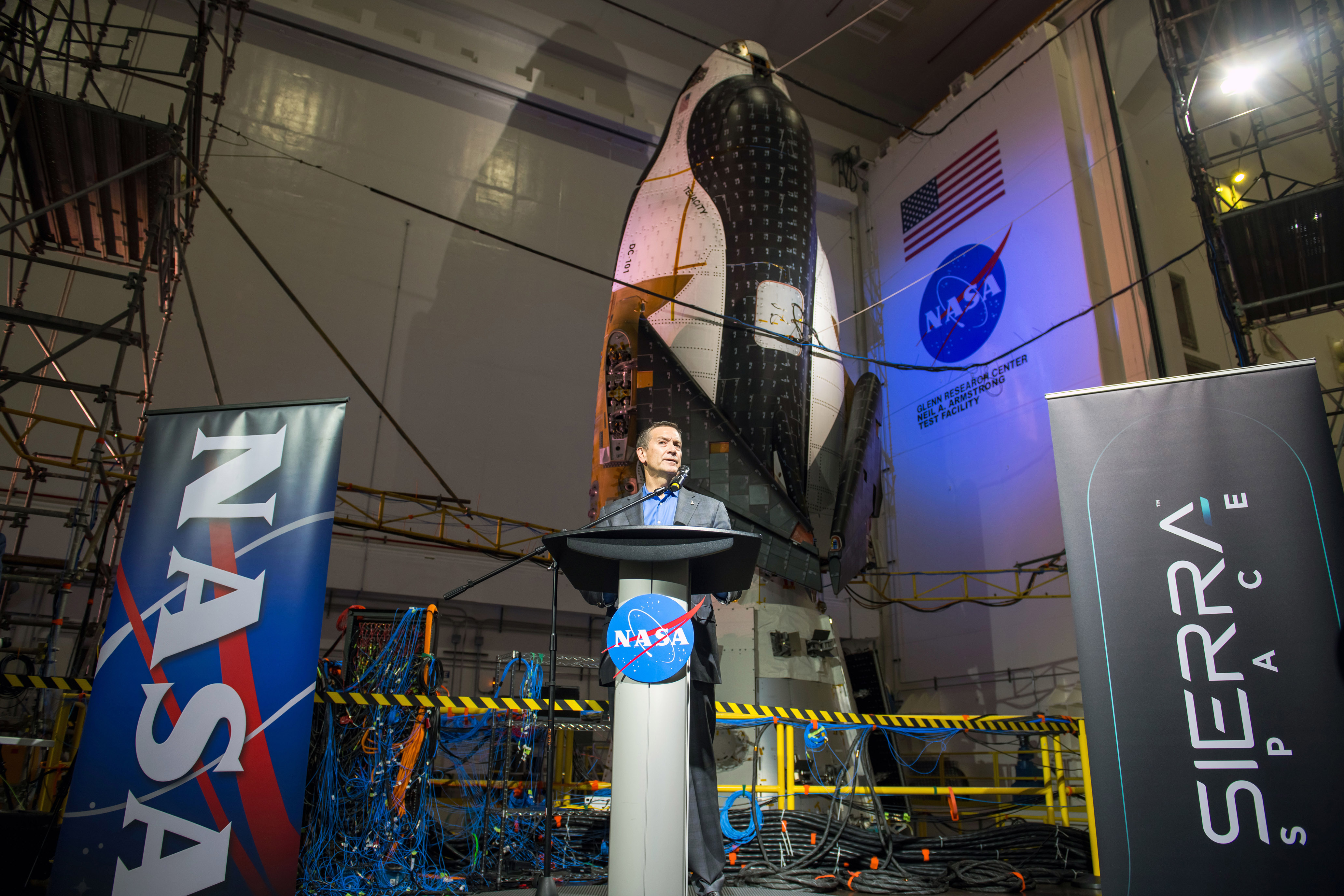 A man in a suit addresses a crowd of media inside the Mechanical Vibration Facility at NASA’s Neil Armstrong Test Facility. He stands at a podium that has a NASA meatball on it. On either side of him are banners that say “NASA” and “Sierra Space.” Behind him is the Dream Chaser spaceplane and its Shooting Star cargo module, nose-up and bathed in soft blue lights. Around the spacecraft are scaffolding and wiring, and a NASA meatball and an American flag can be seen on the wall.