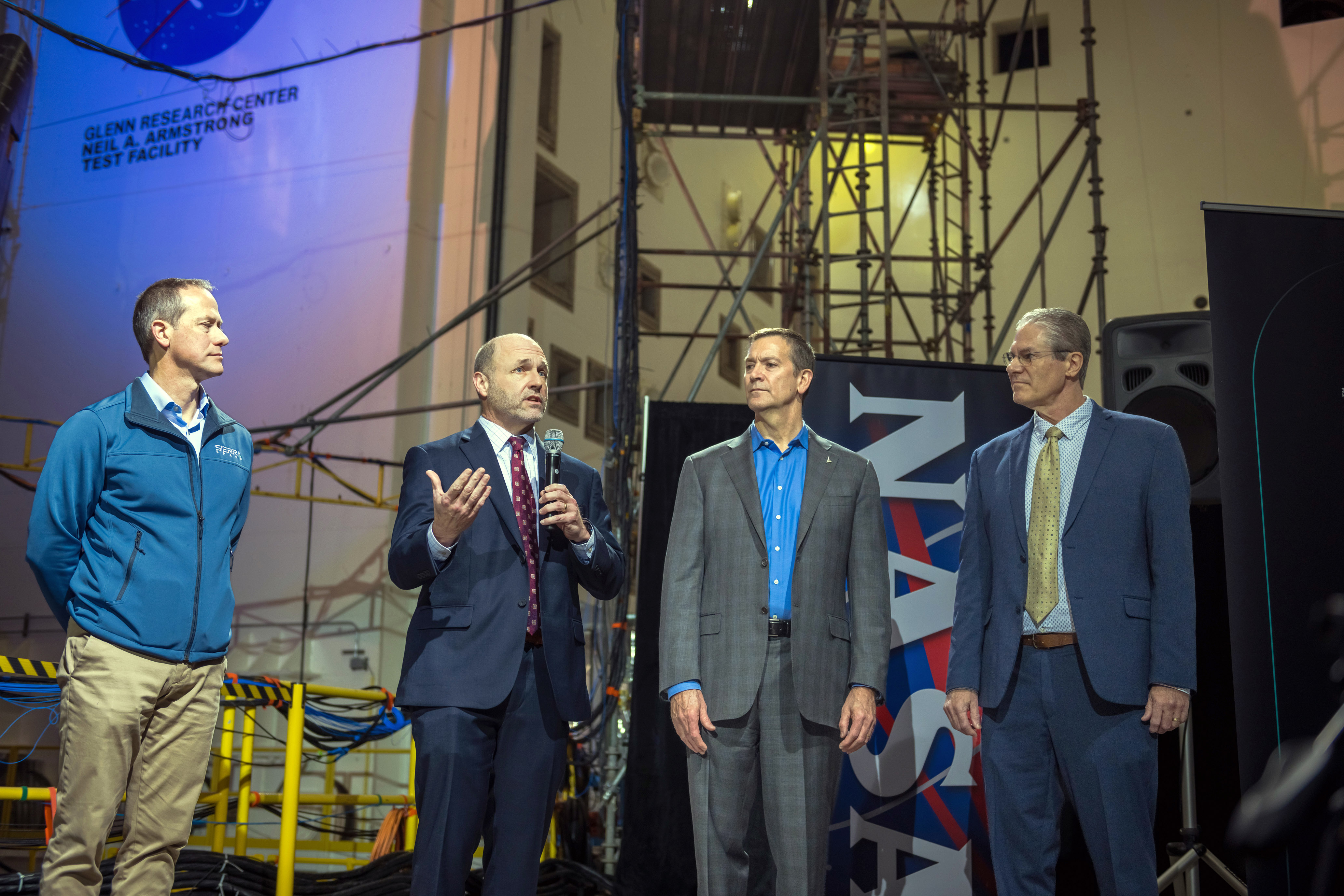 Four men stand inside the Mechanical Vibration Facility at NASA’s Neil Armstrong Test Facility. The three on the right are dressed in suits, and one to the far left is wearing khakis and a jacket that has a Sierra Space logo. They stand in front of a NASA banner, and one holds a microphone to address a crowd of media.