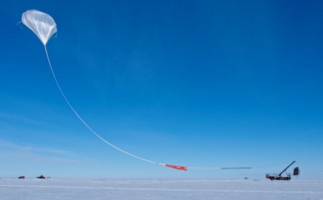 A large white balloon floats into a clear blue sky above snow covered land. Long white material attaches it to a crane holding a scientific payload in the air. It has large black solar panel squares on the sides.