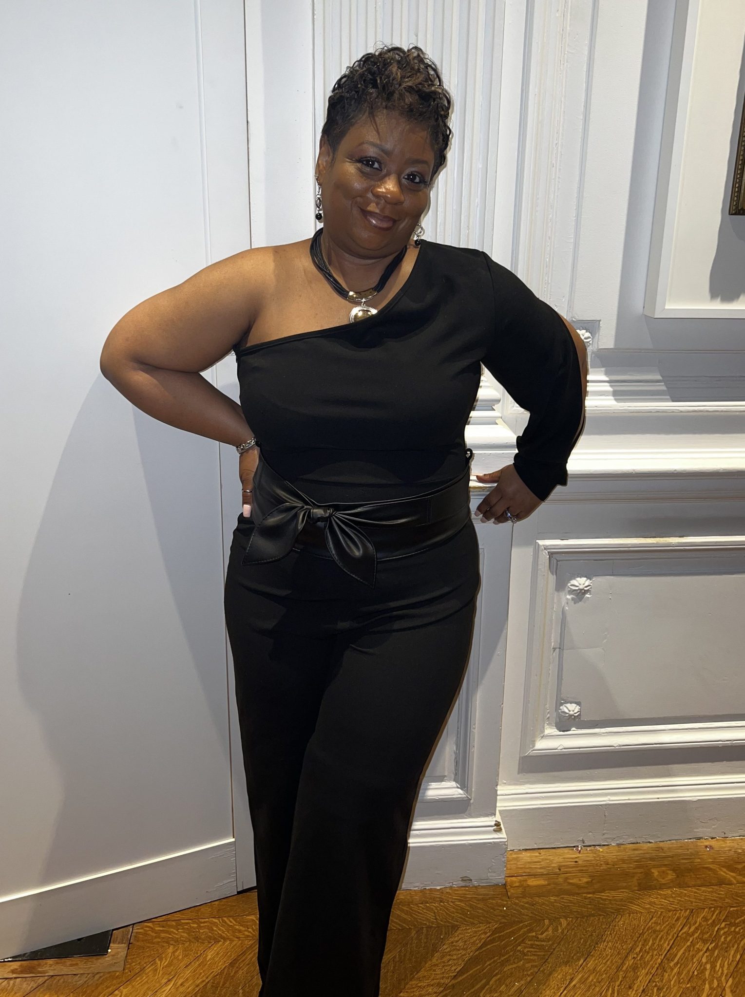 Renee King stands against a white wall wearing a dressy black shirt and black pants. 