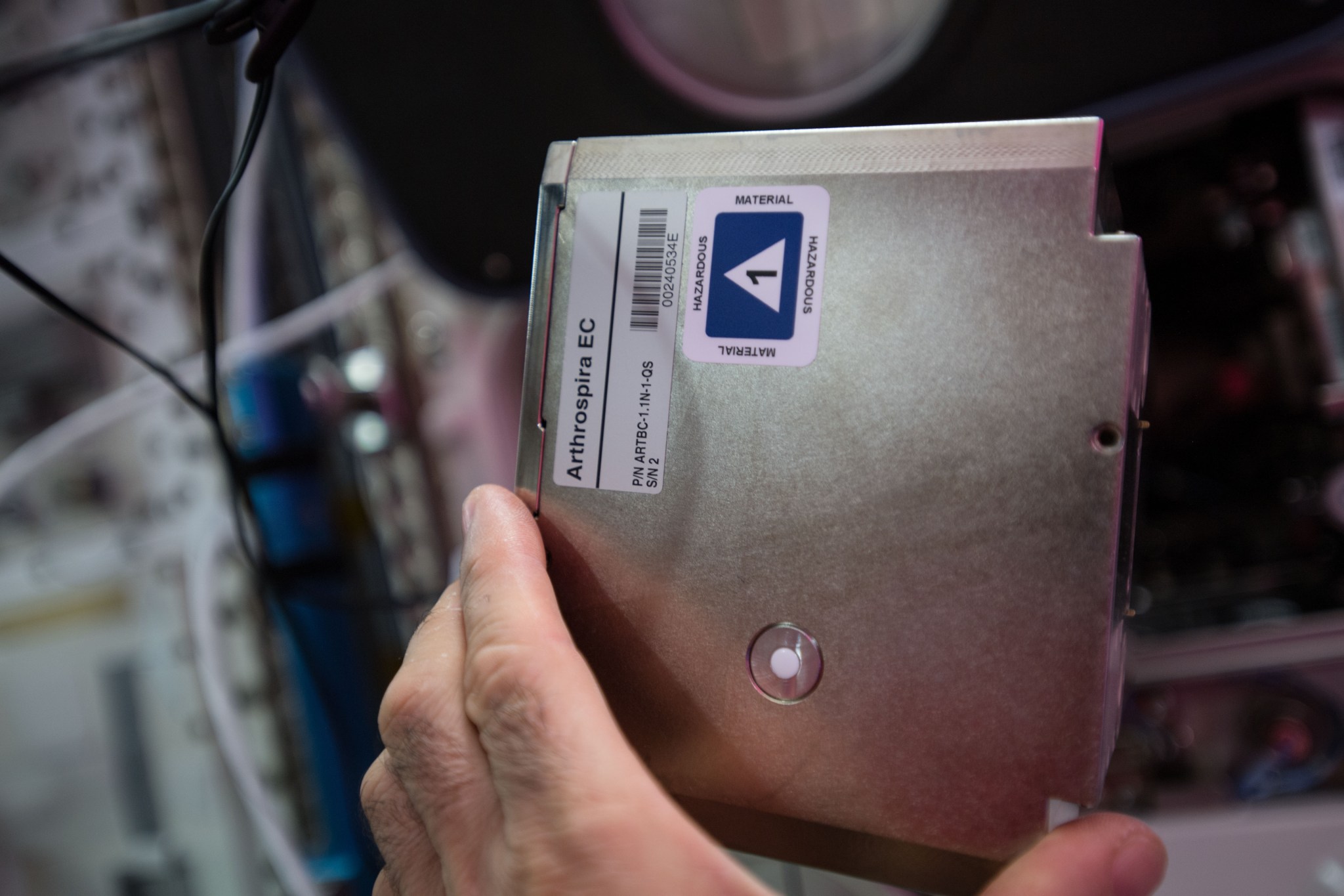 A hand holds a square silver device about the size of a paperback book with a button in the middle of its bottom half and two labels in the upper left corner.