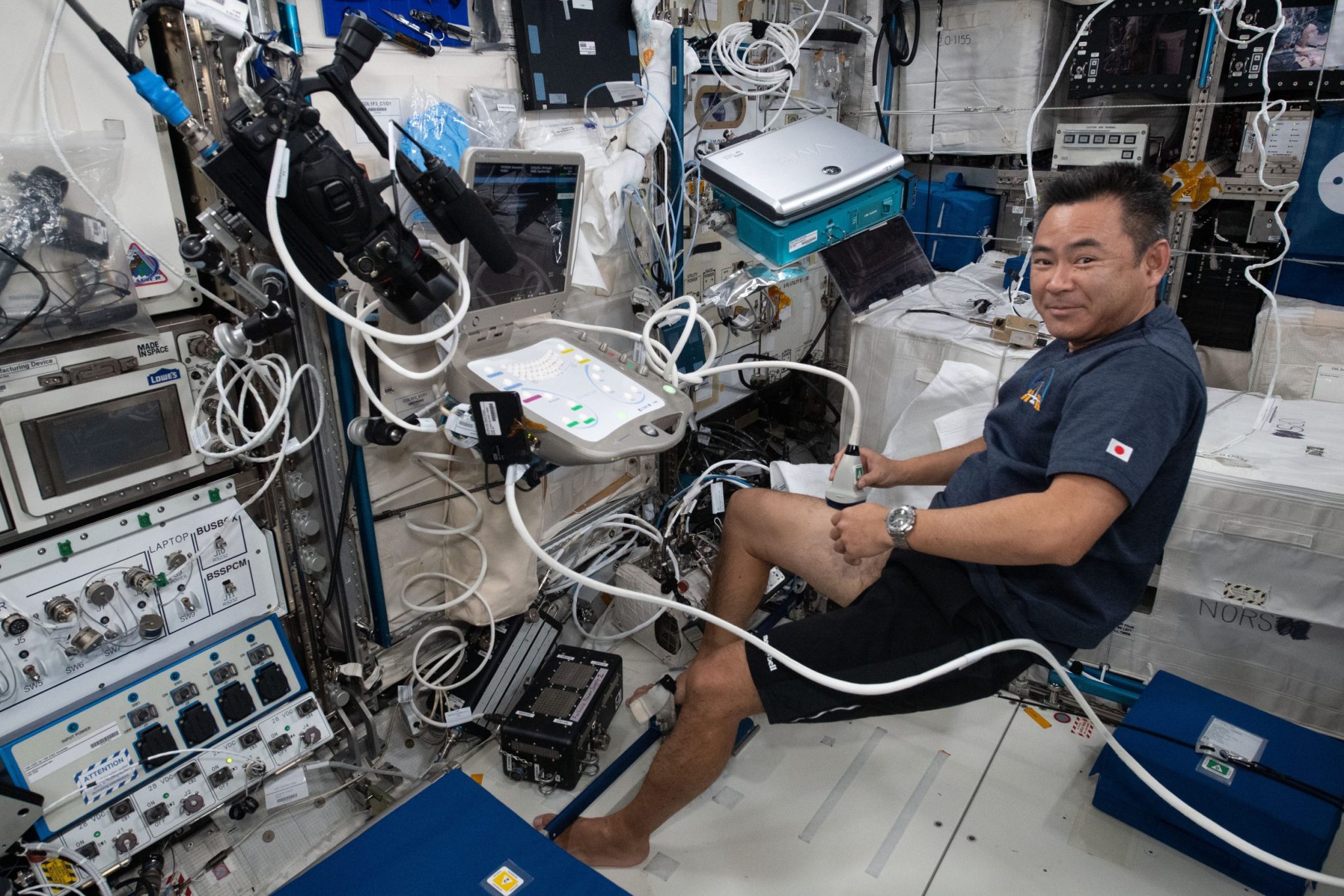Hoshide wears a blue shirt and black short. He holds a small white device, attached by a white cord to a control panel, against his thigh and looks at the camera. The wall in front of him is a jumble of cords, wires, and equipment.