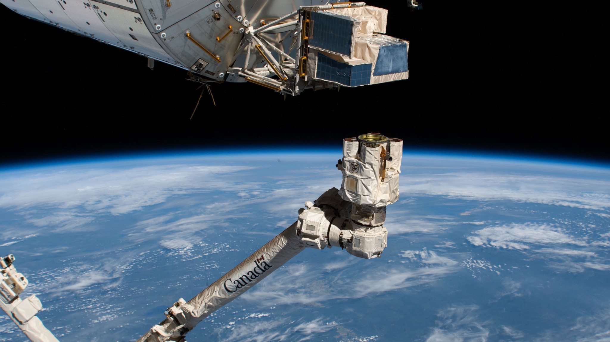 A long white robotic arm extends up from the bottom of the image with “Canada” printed in large letters on its side and sensors showing on the end. Above the arm, ASIM has white protective coverings around blue instruments. Below is the blue Earth with some thin scattered clouds.