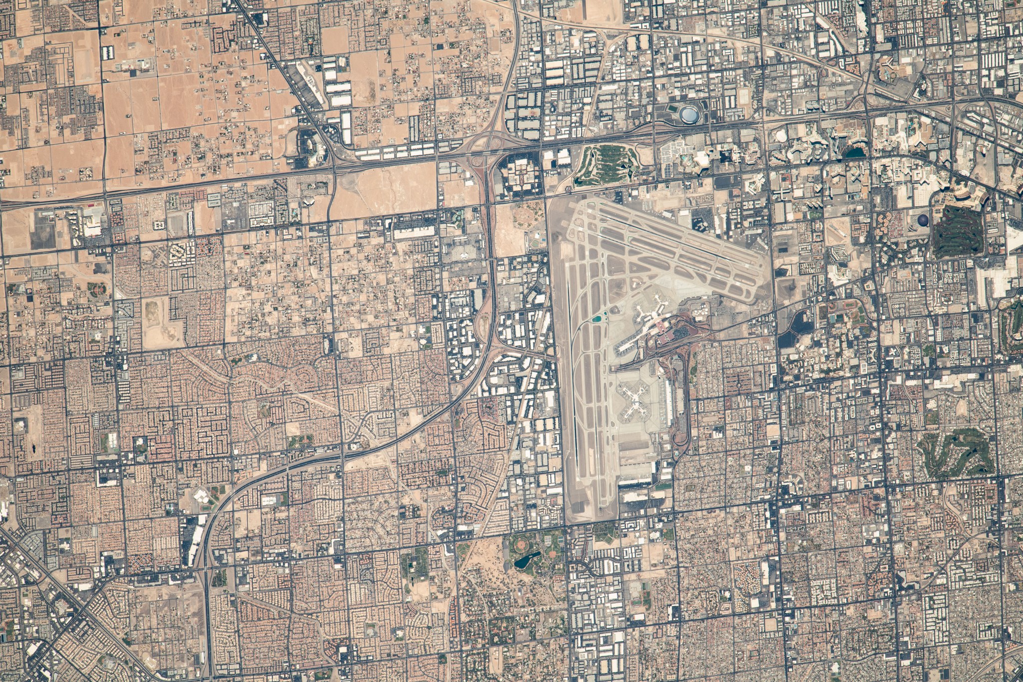 A view of Las Vegas from space. Harry Reid International Airport is visible slightly to the right of the center of the image, and Allegiant Stadium is visible just above the airport. The land is a variety of tans and greens, and there are many square city patterns surrounding the airport on all sides.
