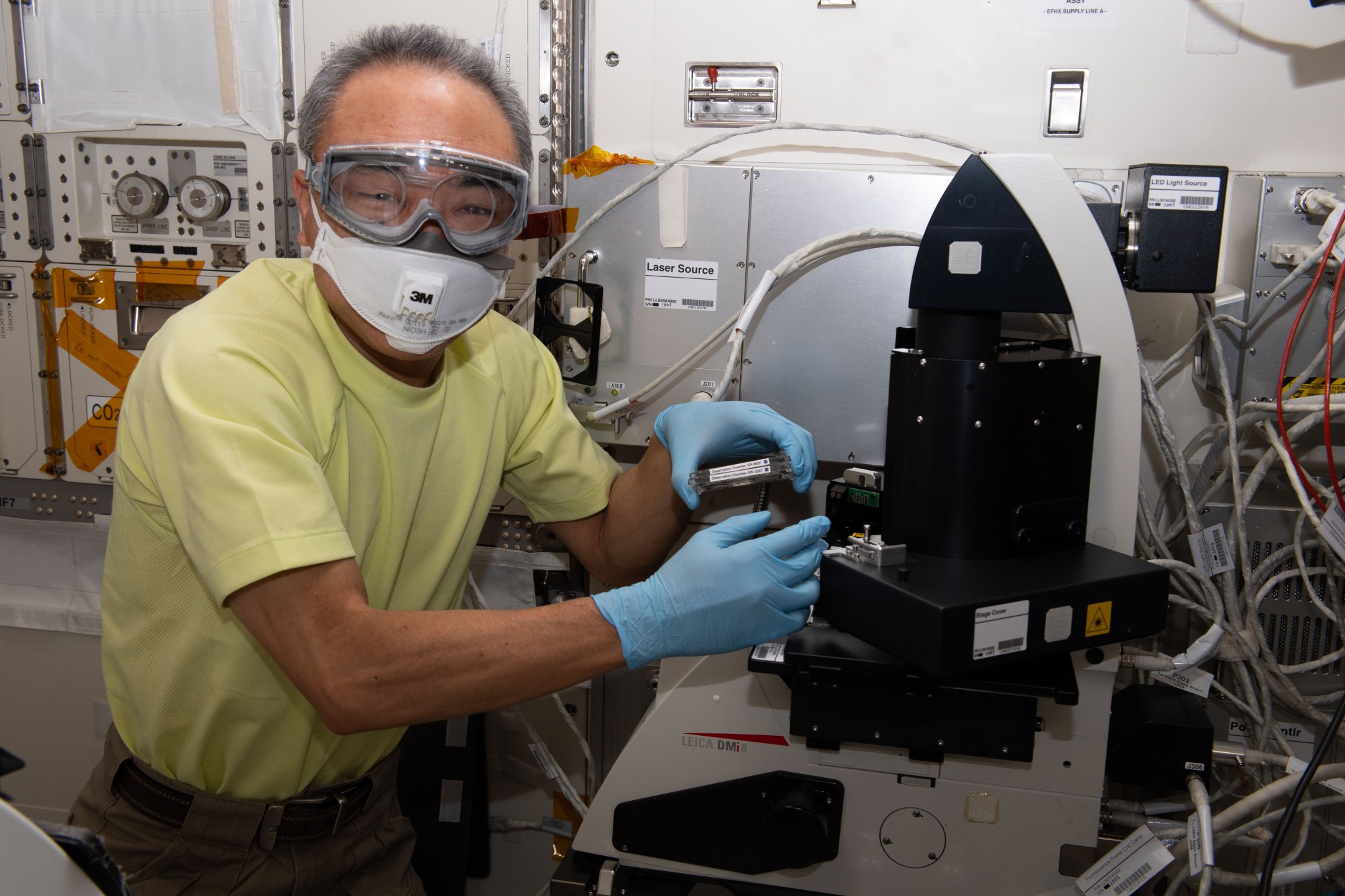 Satoshi Furukawa is wearing a yellow short-sleeved shirt, a mask, googles, and blue gloves as he works with a black microscope on a workbench.